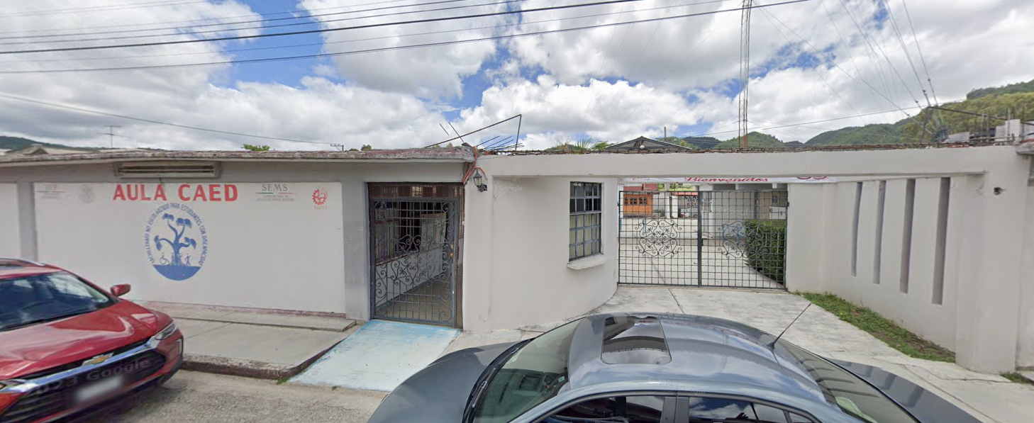 Fotografía en la que se muestra la entrada del plantel conformada por un muro blanco que dice en letras grandes rojas AULA CAED, debajo del texto está pintado el logo de CAED en color azul. Del lado derecho del muro hay una puerta con una rampa que da acceso al plantel y a lado de la puerta hay un portón metálico. 